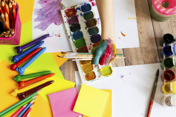 A gyerek fest. Egy gyermek tenyerei és kezei színes akvarellben. Akvarell, gouache, ecsetek, színes és viaszos zsírkréták, matricák. Rajzolás, kreativitás és hobbi. - Fotó, kép