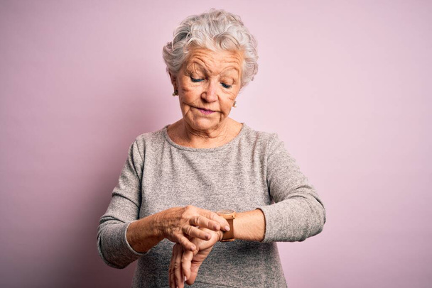 Старша красива жінка в повсякденній футболці стоїть над ізольованим рожевим тлом Перевірка часу на наручні годинники, спокійно і впевнено
 - Фото, зображення