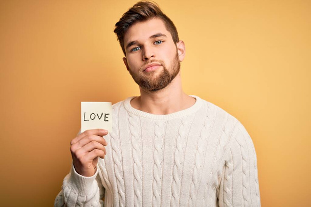 Jovem homem romântico loiro com barba e olhos azuis segurando papel com mensagem de amor com uma expressão confiante no rosto inteligente pensando sério
 - Foto, Imagem