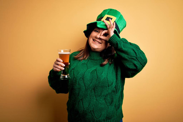 Jovem bonita plus size mulher vestindo chapéu verde celebrando st patricks dia beber cerveja com o rosto feliz sorrindo fazendo ok sinal com a mão no olho olhando através dos dedos
 - Foto, Imagem