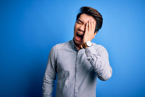 Homem chinês bonito jovem vestindo camisa casual em pé sobre fundo azul isolado bocejo cansado cobrindo metade do rosto, olho e boca com a mão. Cara dói na dor
. - Foto, Imagem