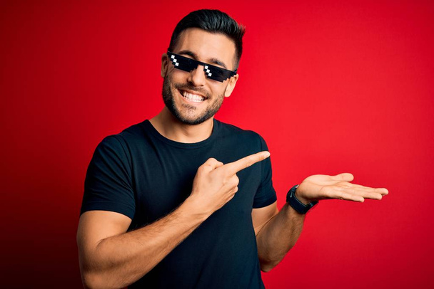 Jonge knappe man draagt grappige schurk leven zonnebril over geïsoleerde rode achtergrond verbaasd en glimlachend naar de camera terwijl presenteren met de hand en wijzen met de vinger. - Foto, afbeelding