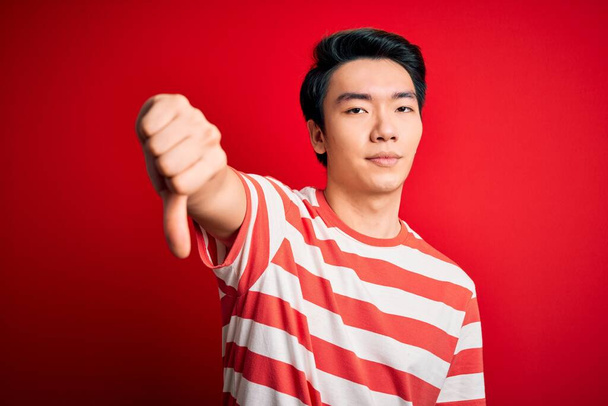 Homem chinês bonito jovem vestindo t-shirt listrada casual em pé sobre fundo vermelho olhando infeliz e irritado mostrando rejeição e negativo com polegares para baixo gesto. Má expressão
. - Foto, Imagem