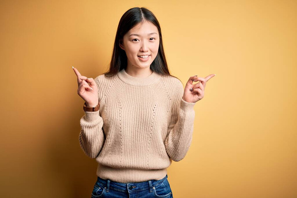 Joven hermosa mujer asiática usando suéter casual sobre fondo aislado amarillo sonriendo confiado señalando con los dedos a diferentes direcciones. Copiar espacio para publicidad
 - Foto, Imagen