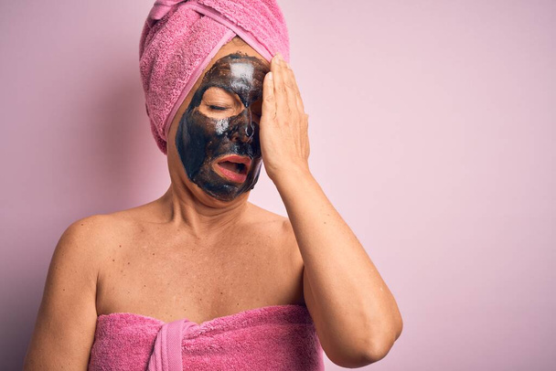 Μεσήλικη μελαχρινή γυναίκα που φοράει μαύρη μάσκα ομορφιάς πάνω από απομονωμένο ροζ φόντο Χασμουρητό κουρασμένο που καλύπτει το μισό πρόσωπο, το μάτι και το στόμα με το χέρι. Το πρόσωπο πονάει από τον πόνο. - Φωτογραφία, εικόνα