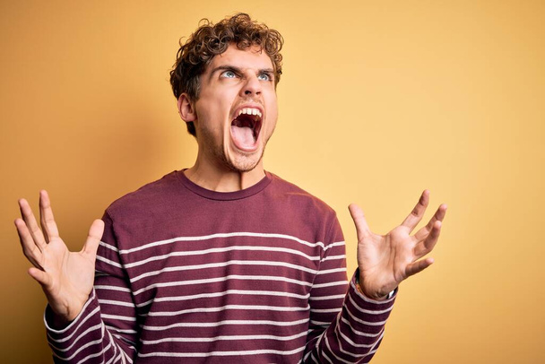 Young blond handsome man with curly hair wearing casual striped sweater crazy and mad shouting and yelling with aggressive expression and arms raised. Frustration concept. - Photo, Image