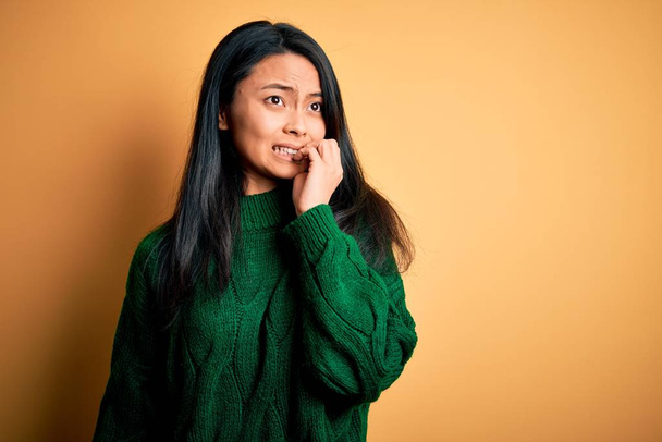 孤立した黄色の背景の上に緑のセーターを着て若い美しい中国の女性は、口の爪をかむ上で手で強調し、緊張している。不安の問題. - 写真・画像