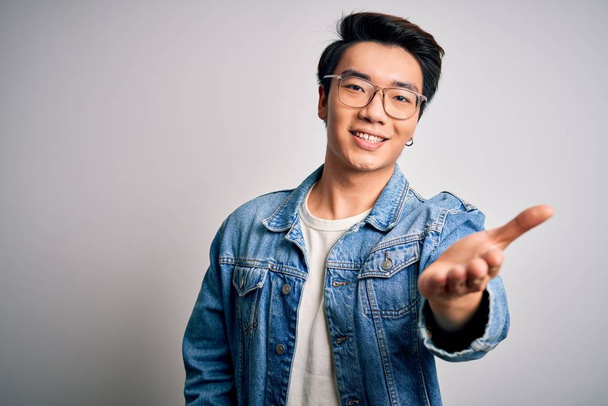 Young handsome chinese man wearing denim jacket and glasses over white background smiling friendly offering handshake as greeting and welcoming. Successful business. - Фото, зображення