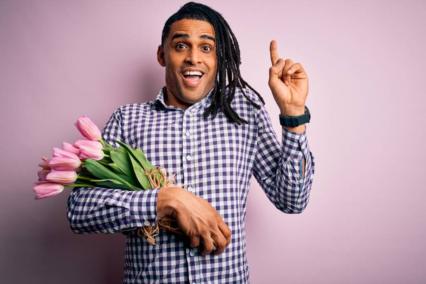 Fiatal afro-amerikai romantikus férfi raszta tartásával csokor rózsaszín tulipán mosolyog csodálkozva és meglepve, és rámutatva ujjaival és felemelt karokkal. - Fotó, kép