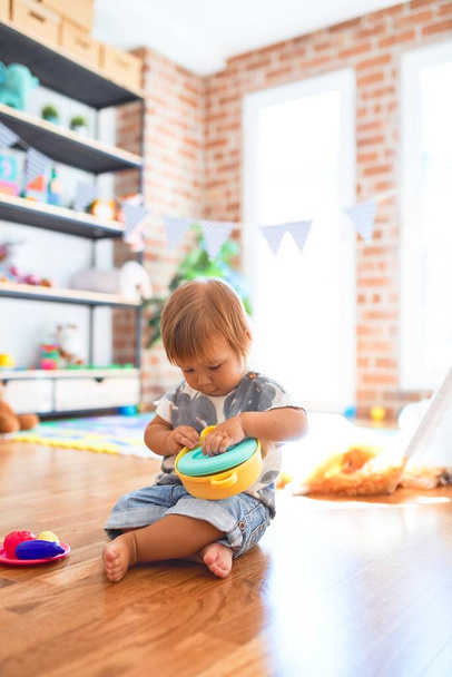 Niño adorable sentado en el suelo jugando comidas usando cubiertos de plástico y comida alrededor de muchos juguetes en el jardín de infantes
 - Foto, imagen