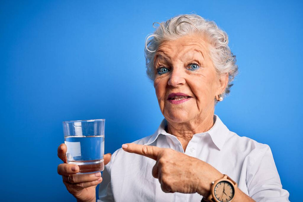 Kıdemli güzel kadın bir bardak su içiyor. Mavi arka planda duruyor. Elinle ve parmağınla işaret ederek çok mutlu. - Fotoğraf, Görsel
