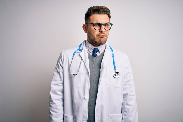 Молодий лікар з блакитними очима носить медичне пальто і стетоскоп над ізольованим тлом скептично і нервово, розчаровуючись через проблеми. Негативна людина
. - Фото, зображення