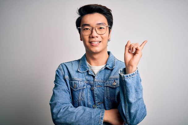 Young handsome chinese man wearing denim jacket and glasses over white background with a big smile on face, pointing with hand and finger to the side looking at the camera. - Φωτογραφία, εικόνα