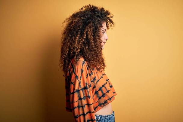 Young beautiful brunette woman with curly hair and piercing wearing casual t-shirt looking to side, relax profile pose with natural face with confident smile. - 写真・画像