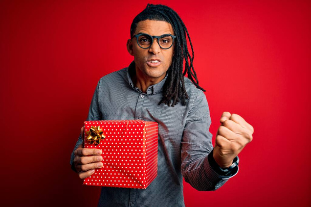 African American man with dreadlocks holding present box over red isolated background annoyed and frustrated shouting with anger, crazy and yelling with raised hand, anger concept - Photo, Image