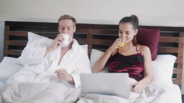 beautiful couple with laptop drinks juice resting in bed - Кадры, видео