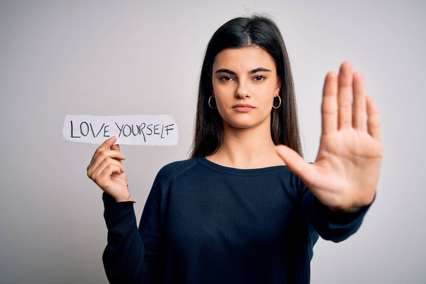 Young beautiful woman asking for take care of you holding paper with love yourself message with open hand doing stop sign with serious and confident expression, defense gesture - Zdjęcie, obraz