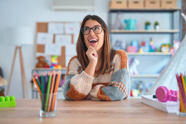 Young beautiful teacher woman wearing sweater and glasses sitting on desk at kindergarten with hand on chin thinking about question, pensive expression. Smiling and thoughtful face. Doubt concept. - Foto, Bild