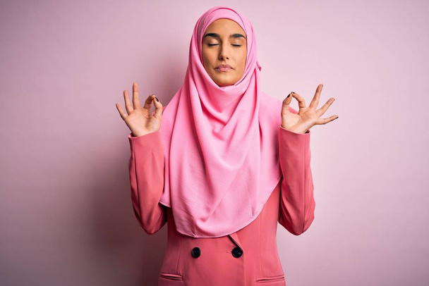 Mladá krásná brunetka podnikatelka v růžové muslimské hidžáb a obchodní bunda relaxovat a usmívat se se zavřenýma očima dělat meditační gesto s prsty. Koncept jógy. - Fotografie, Obrázek