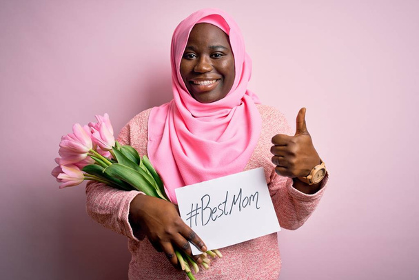 Además de tamaño mujer afroamericana usando hijab sosteniendo mensaje y tulipanes en el día de las madres feliz con una gran sonrisa haciendo signo ok, pulgar hacia arriba con los dedos, señal excelente
 - Foto, imagen