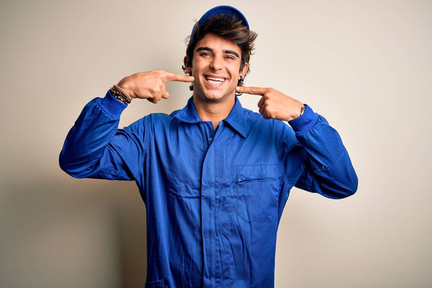 Young mechanic man wearing blue cap and uniform standing over isolated white background smiling cheerful showing and pointing with fingers teeth and mouth. Dental health concept. - Photo, Image