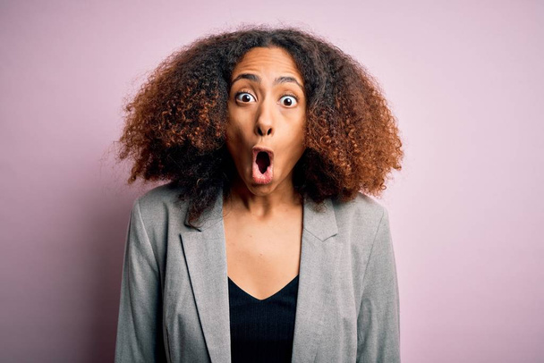 Jeune femme d'affaires afro-américaine aux cheveux afro portant une veste élégante effrayée et choquée par l'expression surprise, la peur et le visage excité
. - Photo, image