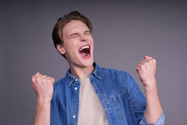 Passionate football fan. A young man in a denim shirt screaming while his team wins, raised his fist in a victory gesture, isolated on a gray background. Hooray, victory. - Photo, Image
