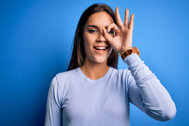 Молодая красивая брюнетка женщина в обычном свитере стоя на синем фоне делает хорошо жест с улыбкой на руке, глаза смотрят сквозь пальцы с счастливым лицом
. - Фото, изображение