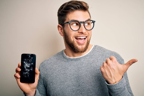 Mladý blonďatý muž s vousy a modrýma očima nosí brýle, drží rozbitý smartphone ukazuje palcem nahoru na stranu se šťastnou tváří usmívá - Fotografie, Obrázek
