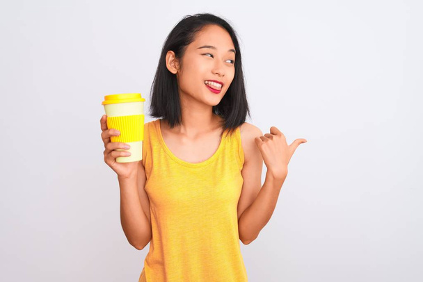 Young chinese woman drinking take away glass of coffee over isolated white background pointing and showing with thumb up to the side with happy face smiling - Photo, Image