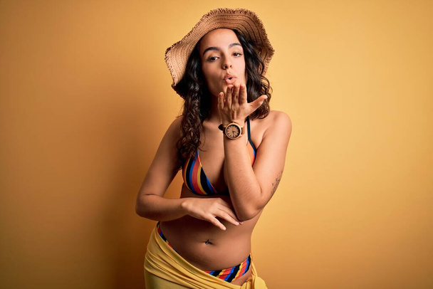 Young beautiful woman with curly hair on vacation wearing bikini and summer hat looking at the camera blowing a kiss with hand on air being lovely and sexy. Love expression. - Foto, immagini