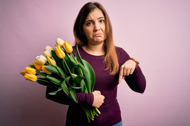Young blonde woman holding romantic bouquet of yellow tulips flowers over pink background Pointing down looking sad and upset, indicating direction with fingers, unhappy and depressed. - Photo, Image