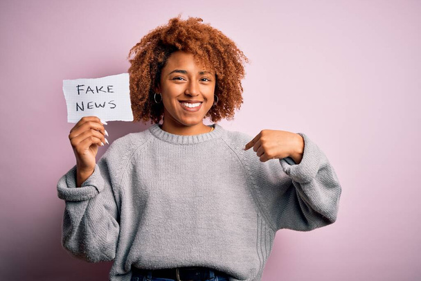 Fiatal afro-amerikai nő göndör haj tartó papír hamis hírek üzenet meglepett arccal mutogatva magára - Fotó, kép