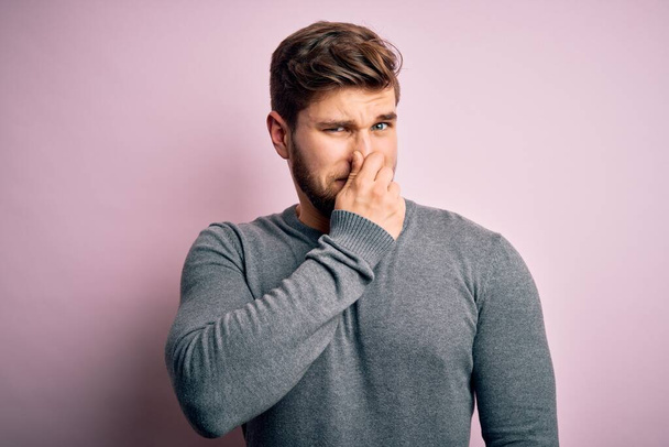Young handsome blond man with beard and blue eyes wearing casual sweater smelling something stinky and disgusting, intolerable smell, holding breath with fingers on nose. Bad smell - Fotó, kép