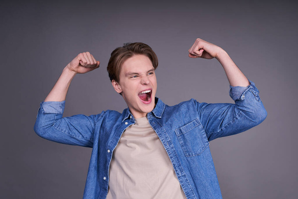 Young man shouting while his team win, raised both fist in victory gesture, isolated on gray background. Casual soccer fan concept. - Foto, afbeelding