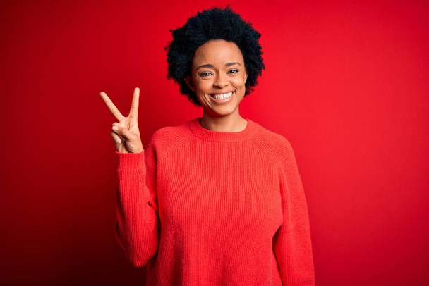 Junge schöne afroamerikanische Afrofrau mit lockigem Haar trägt lässigen Pullover und zeigt mit Fingern Nummer zwei nach oben, während sie selbstbewusst und glücklich lächelt. - Foto, Bild