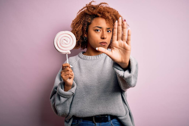 Jovem afro mulher afro afro afro bonita afro com cabelo encaracolado comer doces doces com a mão aberta fazendo sinal de parada com expressão séria e confiante, gesto de defesa
 - Foto, Imagem