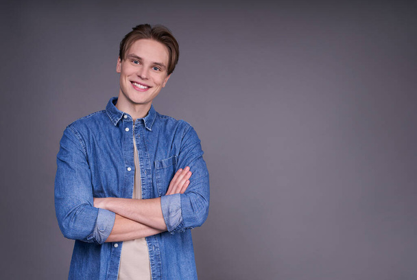 An ambitious guy in a jeans shirt smiling, looking directly with arms crossed, as if planning a business development strategy, isolated on a gray background. Copy space. - Valokuva, kuva