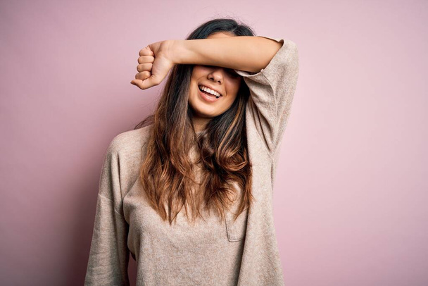 Młoda piękna brunetka ubrana w luźny sweter stojąca nad różowym tłem pokrywająca oczy uśmiechniętym ramieniem wesołym i zabawnym. Koncepcja niewidomych. - Zdjęcie, obraz