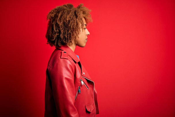 Jeune belle afro afro-américaine aux cheveux bouclés portant une veste rouge décontractée regardant vers le côté, pose de profil relax avec un visage naturel avec un sourire confiant
. - Photo, image