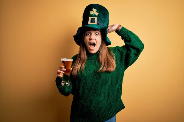 Young beautiful woman wearing green hat drinking glass of beer on saint patricks day Crazy and scared with hands on head, afraid and surprised of shock with open mouth - Фото, зображення