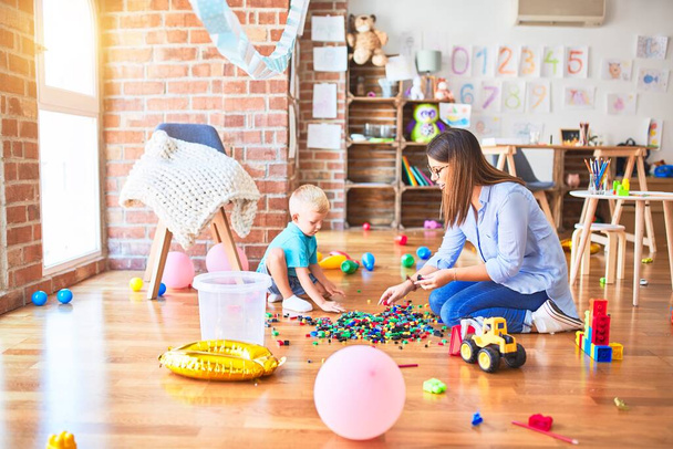 Νεαρό καυκάσιο παιδί που παίζει στο σχολείο με δάσκαλο. Μητέρα και γιος στο playroom φουσκώνουν έναν πύργο με τουβλάκια παιχνιδιών - Φωτογραφία, εικόνα