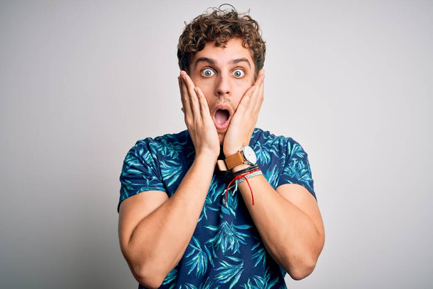 Young blond handsome man on vacation with curly hair wearing casual summer t-shirt afraid and shocked, surprise and amazed expression with hands on face - Photo, Image