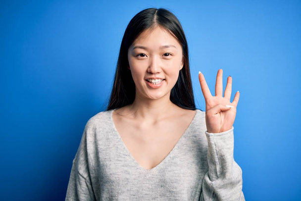 Fiatal, gyönyörű ázsiai nő, alkalmi pulóvert visel, kék, elszigetelt háttér felett áll, mutatva és mutatva a negyedik ujjal, miközben magabiztosan és boldogan mosolyog.. - Fotó, kép