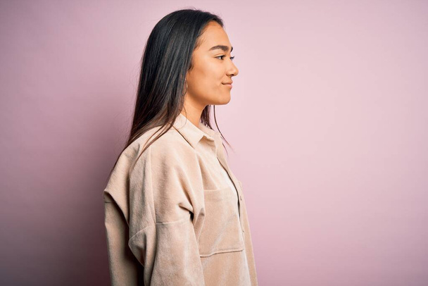 Junge schöne asiatische Frau trägt lässiges Hemd, das über rosa Hintergrund steht und zur Seite schaut, entspannte Profil-Pose mit natürlichem Gesicht und selbstbewusstem Lächeln. - Foto, Bild