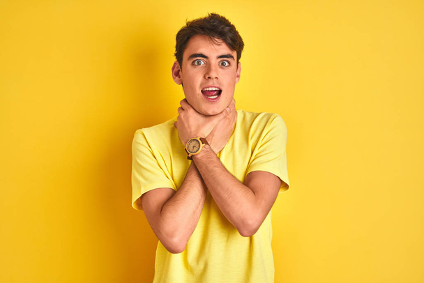 Ένα έφηβο αγόρι που φοράει κίτρινο μπλουζάκι πάνω από απομονωμένο φόντο φωνάζοντας και ασφυκτιώντας επειδή πονάει ο στραγγαλισμός. Πρόβλημα υγείας. Έννοια ασφυξίας και αυτοκτονίας. - Φωτογραφία, εικόνα