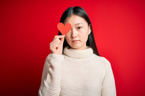 Fiatal ázsiai nő kezében romantikus piros szív papír alakja felett piros elszigetelt háttér magabiztos kifejezés okos arc gondolkodás komoly - Fotó, kép