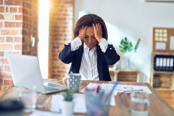 Middelbare leeftijd mooie zakenvrouw werken met behulp van laptop op kantoor lijdt aan hoofdpijn wanhopig en gestrest omdat pijn en migraine. Handen op het hoofd. - Foto, afbeelding