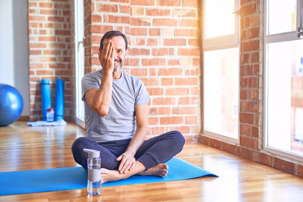 Middelbare leeftijd knappe sportman zittend op mat doen stretching yoga oefening in de sportschool bedekken van een oog met de hand, zelfverzekerde glimlach op het gezicht en verrassing emotie. - Foto, afbeelding