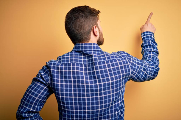 Young blond businessman with beard and blue eyes wearing shirt over yellow background Posing backwards pointing ahead with finger hand - Photo, Image